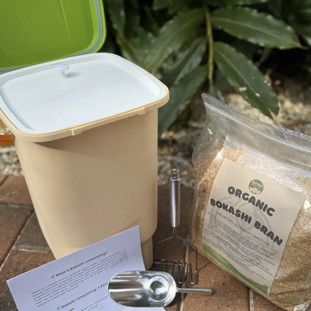 Beige compost bin with green lid, organic bokashi bran package, and metal scoop on a brick surface.